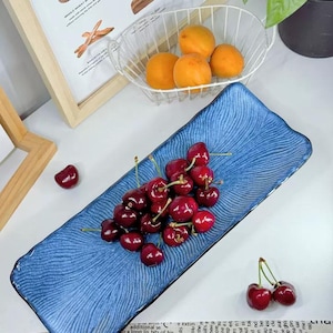 May include: A rectangular blue ceramic serving dish with a textured wave pattern, filled with fresh red cherries. Apricots are in a wire basket in the background. A framed print of bread types is also visible.