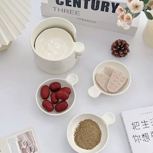 Può includere: Una collezione di piccole ciotole in ceramica bianca con un caratteristico design con manico. Alcune ciotole contengono datteri rossi, cibo a fette e una polvere marrone. Sono presenti anche una ciotola più grande e una tazza, insieme a un libro e una pigna decorativa.
