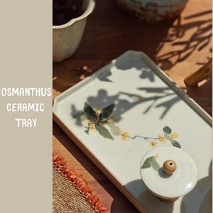 May include: A rectangular ceramic tray with a floral design and the text "OSMANTHUS CERAMIC TRAY". The tray is off-white with a light brown border. The floral design features green leaves and yellow flowers. A matching lid with a wooden knob is also visible.