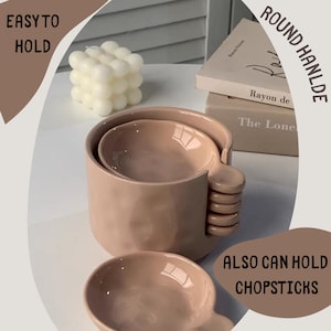 Può includere: Un set di tre ciotole in ceramica color rosa cipria con un design unico del manico. Le ciotole sono annidate l'una nell'altra, con la ciotola più grande con un manico con elementi arrotondati impilati. Il testo "EASY TO HOLD" e "ALSO CAN HOLD CHOPSTICKS" è visibile.