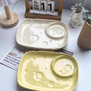 Könnte beinhalten: Zwei rechteckige Keramikplatten, eine cremefarbene und eine gelbe, jeweils mit einer kreisförmigen Vertiefung und mondförmigen Aussparungen. Ein Holz-Kalender zeigt "SEP 11".