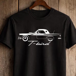 Puede incluir: Camiseta negra con un gráfico blanco de un coche clásico, un Ford Thunderbird, y el texto en cursiva "T-bird". La camiseta está colgada en una percha de madera sobre un fondo de madera.