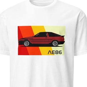 May include: White t-shirt featuring a red sports car design with a black roof and the text "AE86" in yellow. The car is set against a backdrop of red, orange, and yellow stripes. A classic car design.