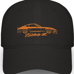 Puede incluir: Gorra de béisbol negra con una ilustración naranja de un coche deportivo Fairlady Z clásico. El diseño muestra la silueta del coche, incluyendo ruedas y detalles de la carrocería. El texto "Fairlady Z" está impreso debajo en naranja.