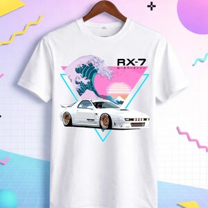 May include: White t-shirt featuring a white sports car graphic with gold rims, set against a pink and teal geometric design with a wave and sunset. The text "RX-7" is displayed above the car.