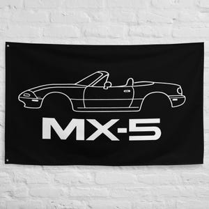 May include: Black flag featuring a white outline of a convertible car. The car's design is a side profile with the text "MX-5" in bold white letters below the car. The flag is hanging against a white brick wall.