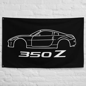 May include: Black flag featuring a white outline of a 350Z sports car. The text "350 Z" is printed in white below the car's image. The flag is rectangular and hangs against a white brick wall.