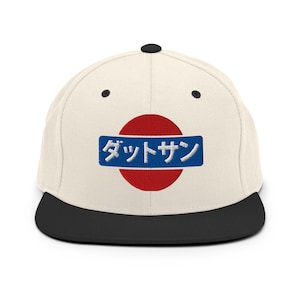 May include: A cream-coloured baseball cap with a black brim and a black button on top. The cap features a red circle with a blue rectangle containing Japanese text.