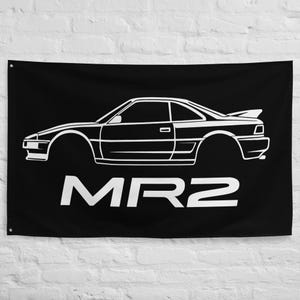 May include: Black flag featuring a white outline of a sports car with a rear spoiler. The word "MR2" is printed in large white block letters below the car. The flag is hanging against a white brick wall.
