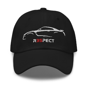 Pode incluir: Boné de beisebol preto com um contorno branco bordado de um carro esportivo. O texto "R35PECT" é bordado em vermelho. O boné tem uma viseira curva e uma alça ajustável.