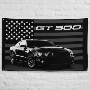 May include: Black and white wall flag featuring a black sports car in front of a grayscale American flag. The text "GT 500" is displayed in white across the top of the flag. The car has a sleek, modern design.