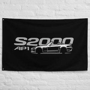 Może przedstawiać: Czarna flaga z białym graficznym przedstawieniem samochodu Honda S2000 AP1. Tekst "S2000 AP1" jest wyświetlany na biało nad sylwetką samochodu. Flaga jest prostokątna i wisi na białej ścianie.