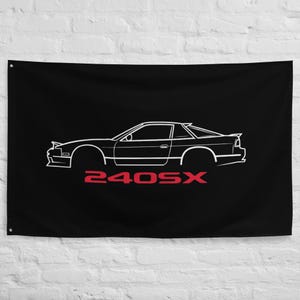 Puede incluir: Bandera negra con un contorno blanco de un coche deportivo y el texto rojo "240SX". El perfil lateral del coche se representa en blanco, destacando su diseño. La bandera está colgada en una pared.