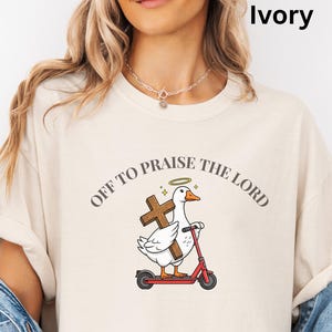 May include: Ivory-colored t-shirt featuring a cartoon goose riding a red scooter and holding a wooden cross. The text "OFF TO PRAISE THE LORD" is arched above the goose. The goose has a halo. The shirt is a light cream color.