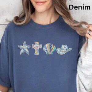 May include: Denim blue t-shirt with a decorative design featuring a starfish, cross, seashell, and cowboy hat. The design is in shades of blue and white. The word "Denim" is printed at the top.