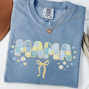 May include: Blue jean color t-shirt with the word "MAMA" in patchwork letters. The shirt has a yellow bow and floral accents. A gold necklace with a pendant is draped across the shirt.