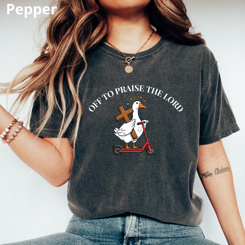 May include: Dark gray t-shirt with the text "OFF TO PRAISE THE LORD". The graphic features a white duck with a halo, holding a cross, riding a red scooter. The shirt is a comfortable, casual style.