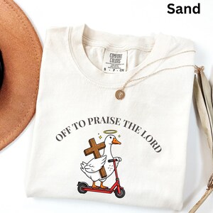 May include: Sand-colored t-shirt with the text "OFF TO PRAISE THE LORD" encircling a cartoon duck riding a red scooter and holding a cross. The shirt has a crew neck and short sleeves. A gold necklace and a brown hat are also visible.
