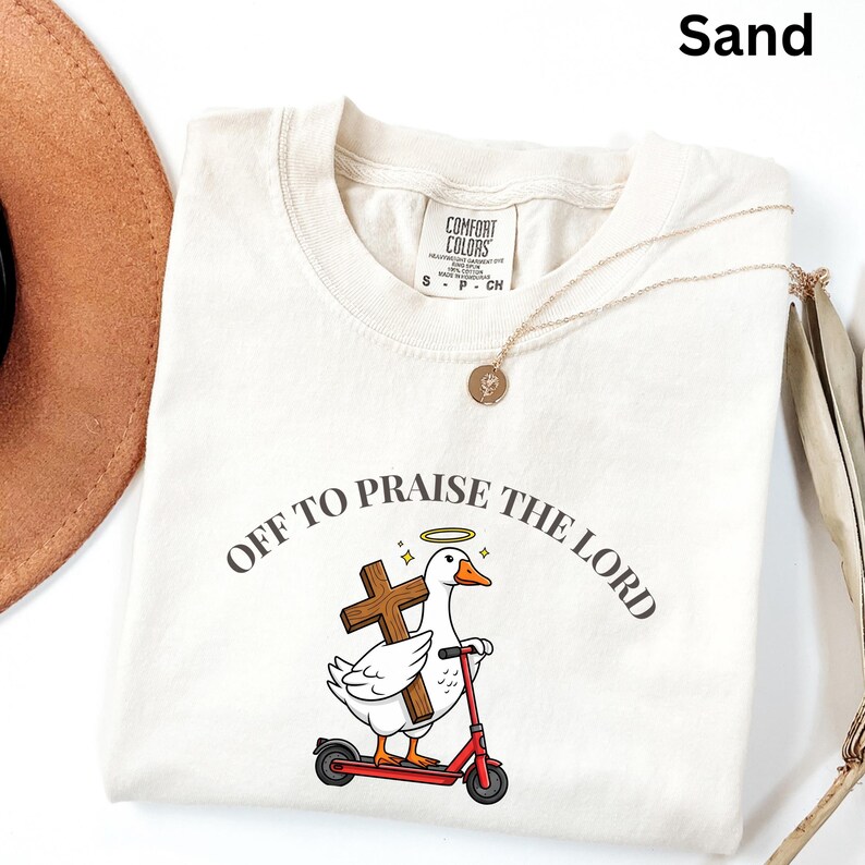 May include: Sand-colored t-shirt with the text "OFF TO PRAISE THE LORD" encircling a cartoon duck riding a red scooter and holding a cross. The shirt has a crew neck and short sleeves. A gold necklace and a brown hat are also visible.