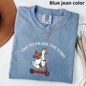 May include: Blue jean-colored t-shirt with the text "OFF TO PRAISE THE LORD." The graphic features a white goose with a halo, holding a cross, riding a red scooter. A gold necklace is draped across the shirt.
