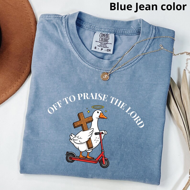 May include: Blue jean-colored t-shirt with the text "OFF TO PRAISE THE LORD." The graphic features a white goose with a halo, holding a cross, riding a red scooter. A gold necklace is draped across the shirt.