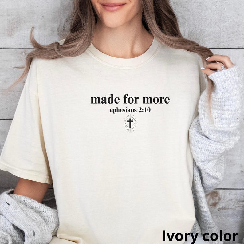 May include: Ivory-colored t-shirt with the text "made for more" above "ephesians 2:10" and a cross graphic. The shirt is a neutral color and has a simple, classic design.