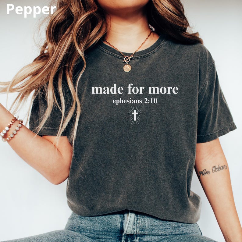 May include: Dark gray t-shirt with the text "made for more" and "ephesians 2:10" in white, along with a cross graphic. The shirt has a crew neck and short sleeves. The shirt is a comfortable, casual style.