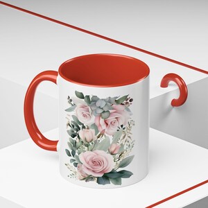 May include: A white ceramic mug with a red rim and handle. The mug features a floral design with pink roses and green leaves.