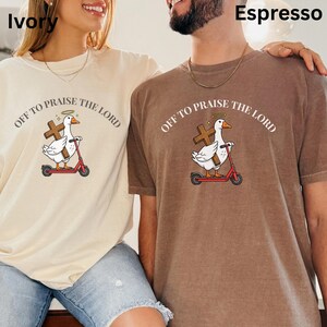 May include: Ivory and espresso-colored t-shirts with the text "OFF TO PRAISE THE LORD" and a graphic of a white duck riding a red scooter and holding a cross. The duck has a halo above its head. The shirts are short-sleeved.