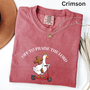 May include: Crimson-colored t-shirt with the text "OFF TO PRAISE THE LORD" and a cartoon goose riding a red scooter while carrying a cross. The shirt has a crew neck and a small tag. A silver necklace with a pendant is also visible.