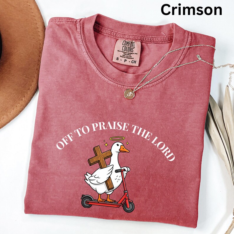 May include: Crimson-colored t-shirt with the text "OFF TO PRAISE THE LORD" and a cartoon goose riding a red scooter while carrying a cross. The shirt has a crew neck and a small tag. A silver necklace with a pendant is also visible.