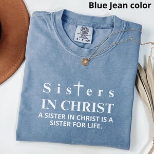 May include: A blue jean colored t-shirt with the text "Sisters in Christ" and "A sister in Christ is a sister for life." The shirt has a distressed look and a small tag that says "Comfort Colors".