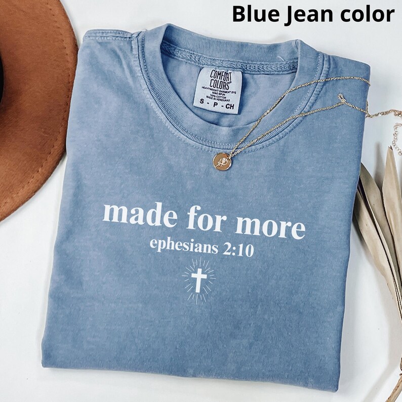 May include: Blue jean color t-shirt with the text "made for more ephesians 2:10" in white. The shirt has a small cross graphic and a gold necklace. The shirt is a Comfort Colors brand, size S-CH.