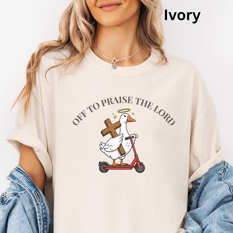 May include: Ivory-colored t-shirt featuring a cartoon goose riding a red scooter and holding a wooden cross. The text "OFF TO PRAISE THE LORD" is arched above the goose. The goose has a halo. The shirt is a light cream color.