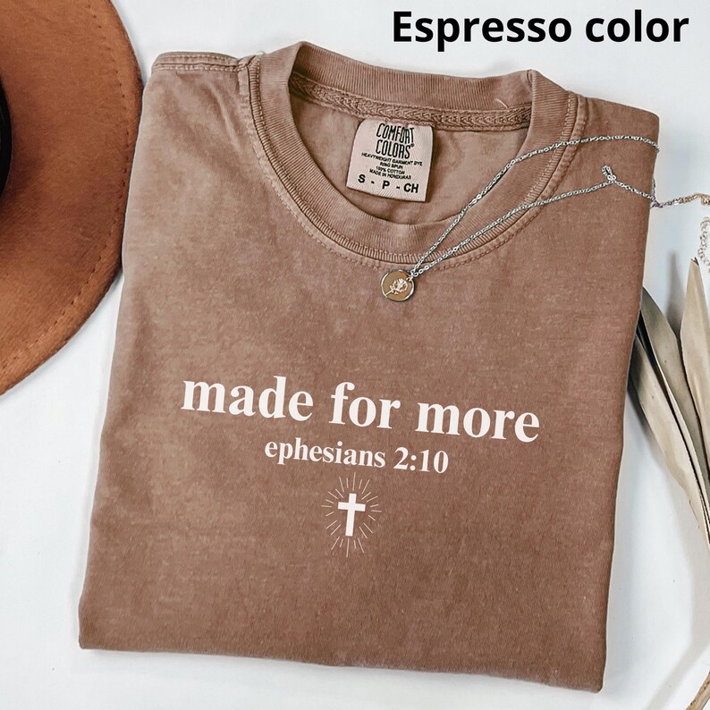 May include: Espresso-colored t-shirt with the text "made for more" and "ephesians 2:10" in white. A silver necklace with a pendant and a cross graphic are also visible. The shirt is a Comfort Colors brand.