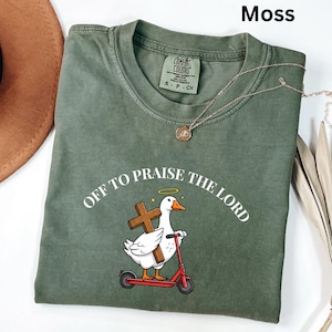 May include: Moss green t-shirt with the text "OFF TO PRAISE THE LORD" around a cartoon goose holding a cross and riding a red scooter. A gold necklace is draped across the shirt. The shirt is made of a soft, comfortable material.