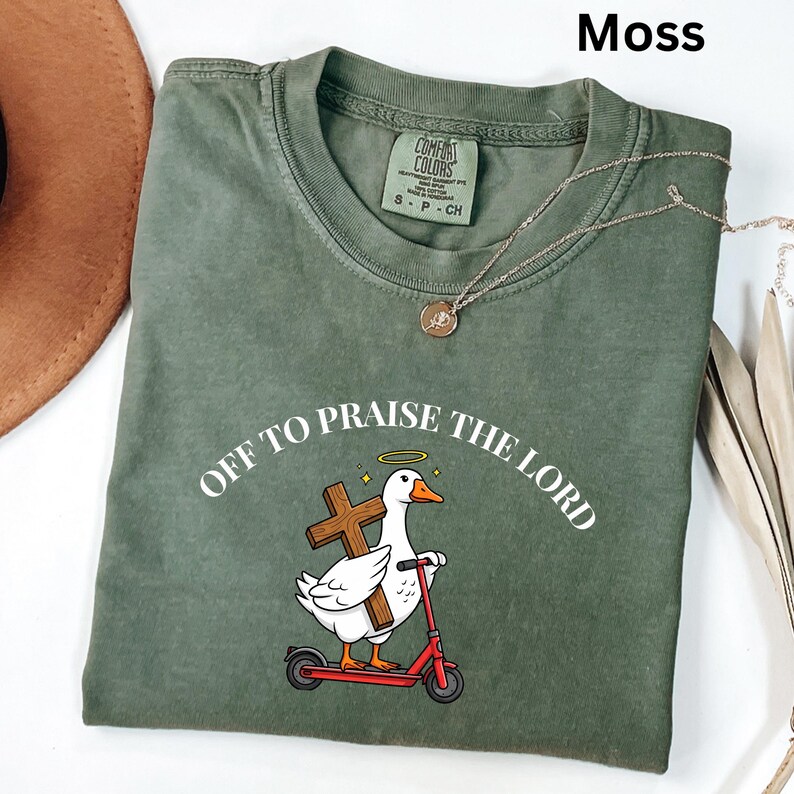 May include: Moss green t-shirt with the text "OFF TO PRAISE THE LORD" around a cartoon goose holding a cross and riding a red scooter. A gold necklace is draped across the shirt. The shirt is made of a soft, comfortable material.