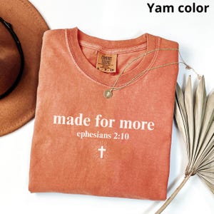 May include: A yam-colored t-shirt with the words "made for more" and the reference "ephesians 2:10" printed in white. A gold necklace with a pendant and a brown hat are also visible.