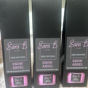 Könnte beinhalten: Drei schwarze Boxen mit weißem Text und rosa Etiketten. Jede Box enthält einen Schilfrohrdiffusor mit der Aufschrift "Sara B Reed Diffuser Snow Angel" und der Website "meltsbysarab.myshopify.com".