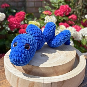 May include: A bright blue crocheted snake toy with a soft, textured yarn finish and two large, dark, glossy plastic eyes. The plush snake is coiled in a spiral shape, showcasing its rounded head and segmented body structure made of yarn.