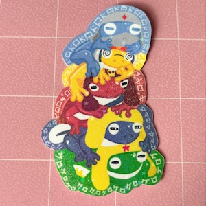May include: Stack of four frog-themed stickers with glitter accents. The top sticker features a blue frog with a red star on its head. The second sticker features a yellow frog with a red star on its head. The third sticker features a red frog with a red star on its head. The bottom sticker features a green frog with a red star on its head. The stickers have Japanese text around the edges.