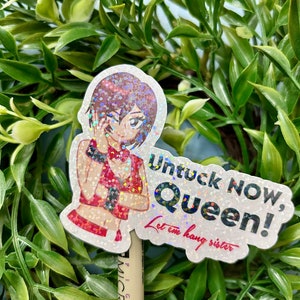 Puede incluir: Una pegatina hologr&aacute;fica con un personaje de estilo anime con un atuendo rojo. La pegatina tiene el texto "Untuck NOW, Queen! Let em hang sister" en una fuente divertida. La pegatina est&aacute; colocada frente a un fondo de hojas verdes.