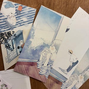 May include: A collection of watercolor art prints featuring serene scenes of people and animals. The prints showcase soft blues and whites, with illustrations of a cat, a person looking out a window, and other figures. The text "生活 Sayuri Fujinaki" is visible on one print.