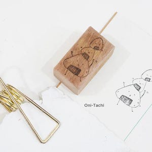 May include: A wooden stamp with a design of a cartoon rice ball character. The stamp is on a white surface with a piece of paper that says "Oni-Tachi" and a drawing of the rice ball character.