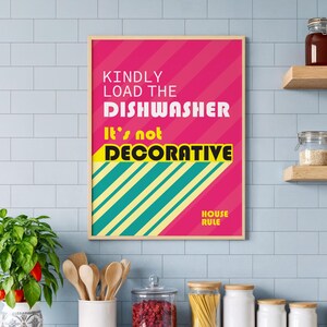 May include: A framed wall art print with the text "KINDLY LOAD THE DISHWASHER It's not DECORATIVE" in white and yellow on a pink background. The print is in a kitchen setting with a plant and jars.