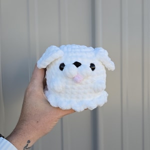 May include: A white crocheted puppy toy with black eyes and a pink tongue. The soft toy has floppy ears and a rounded body. The puppy is held in a hand against a grey background.