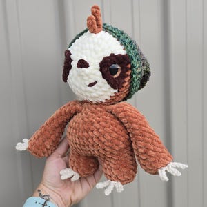 May include: A plush sloth toy made of brown chenille yarn with a cream-colored face, dark brown eye patches, and large black eyes. The sloth wears a textured green and brown beanie hat and has white yarn claws on its hands and feet.