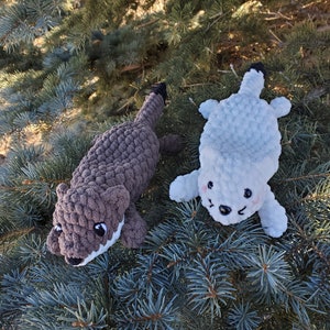 May include: Two crocheted stuffed animals, one brown and one light blue, are posed on a green evergreen tree branch. The brown animal has a white mouth and black eyes. The light blue animal has a white face with black eyes and a pink nose.