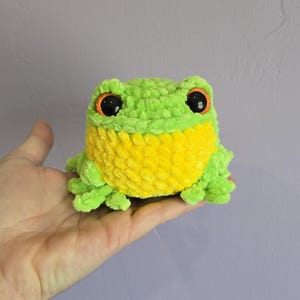 May include: A handmade, plush frog toy in shades of green and yellow. The frog has large, black eyes with orange accents. The soft, textured material gives it a cuddly appearance. A charming, whimsical item.