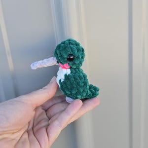 Peut inclure: Un colibri fait à la main au crochet, dans des tons de vert, blanc, rose et gris. L'oiseau a un corps texturé, un long bec gris et de grands yeux noirs. Le colibri est un petit jouet en peluche.